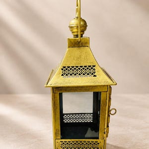 Puede incluir: Una linterna de metal dorado con acabado desgastado, base cuadrada, cuatro paneles de vidrio y recortes en forma de coraz&oacute;n en los bordes superior e inferior. Incluye un asa en la parte superior y un peque&ntilde;o pestillo lateral para abrir.