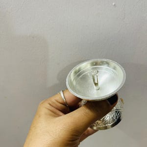 May include: A silver-colored metal oil lamp featuring a wide, shallow bowl with a central wick holder. The exterior of the base is decorated with intricate, embossed floral patterns, providing a textured appearance to the metallic surface.