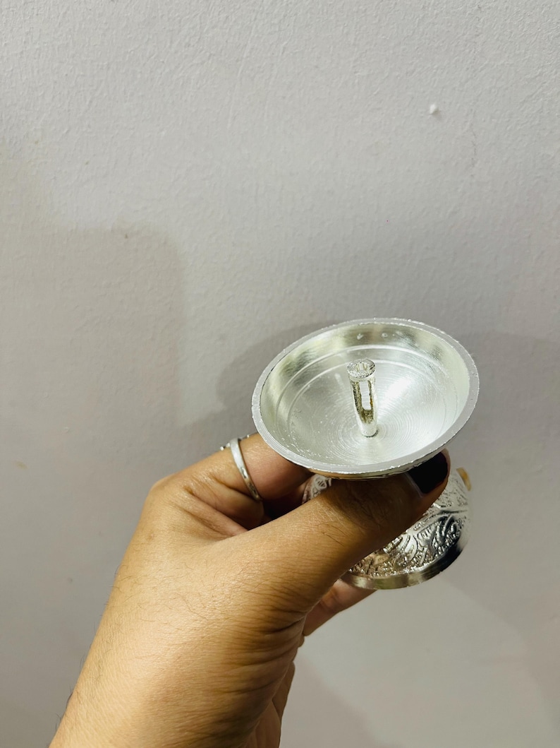 May include: A silver-colored metal oil lamp featuring a wide, shallow bowl with a central wick holder. The exterior of the base is decorated with intricate, embossed floral patterns, providing a textured appearance to the metallic surface.
