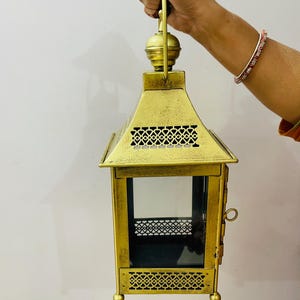 Puede incluir: Un farol de metal dorado con acabado envejecido, que presenta una base cuadrada, cuatro paneles de cristal y complejos patrones geom&eacute;tricos recortados en las secciones superior e inferior. Incluye una puerta lateral y un asa superior.