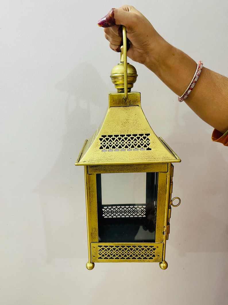 Puede incluir: Un farol de metal dorado con acabado envejecido, que presenta una base cuadrada, cuatro paneles de cristal y complejos patrones geom&eacute;tricos recortados en las secciones superior e inferior. Incluye una puerta lateral y un asa superior.