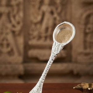 May include: A silver-toned spoon with a textured, shell-like handle and a round bowl containing a light brown powder. The spoon features intricate embossed detailing along the stem and base, with a pointed, tapered end resembling a conch shell.