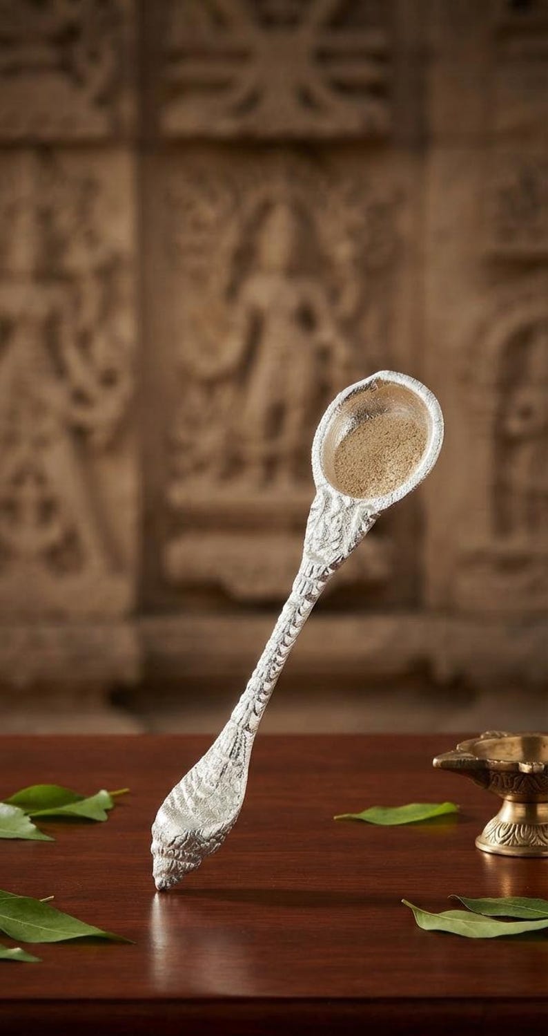 May include: A silver-toned spoon with a textured, shell-like handle and a round bowl containing a light brown powder. The spoon features intricate embossed detailing along the stem and base, with a pointed, tapered end resembling a conch shell.