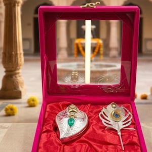 May include: A bright pink velvet display box with a clear lid containing two silver-toned items. On the left is a leaf-shaped silver object with a green teardrop stone. On the right is a silver peacock feather-shaped ornament with a blue and gold accent.