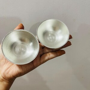 May include: Two small, circular, metallic bowls with a polished silver-toned finish. The bowls feature a smooth, reflective interior surface and a simple, rounded rim design. These lightweight, open-top containers are presented together.