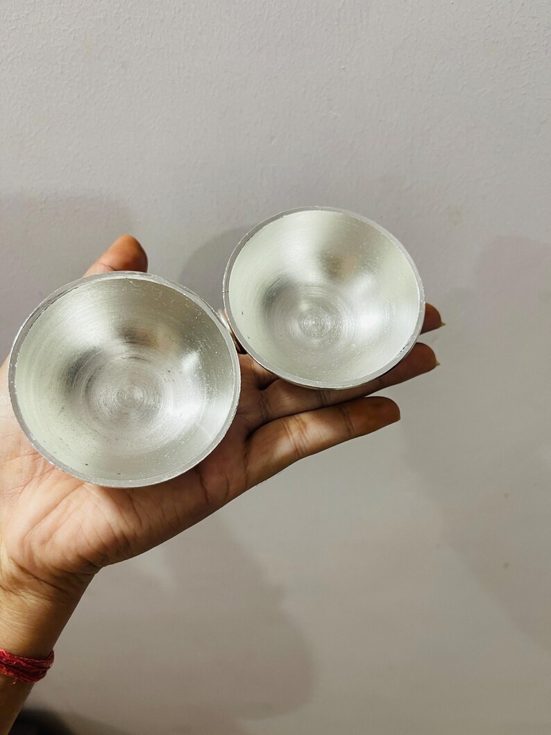 May include: Two small, circular, metallic bowls with a polished silver-toned finish. The bowls feature a smooth, reflective interior surface and a simple, rounded rim design. These lightweight, open-top containers are presented together.