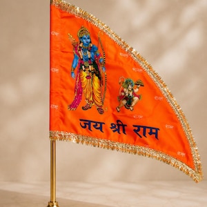 Könnte beinhalten: Eine dreieckige orangefarbene Flagge mit einer blauhäutigen Gottheit, die einen Bogen hält, und einer kleineren affenartigen Figur, mit dem Hindi-Text "जय श्री राम" in Blau. Die Flagge ist mit Goldfransen verziert und an einem goldfarbenen Metallstab befestigt.