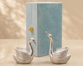 Puede incluir: Dos portavelas plateados con forma de cisne, con detalles de alas texturizadas e inserciones de vidrio transparente, situados frente a una caja de almacenamiento rectangular alta, cubierta de terciopelo azul claro con un cierre dorado.