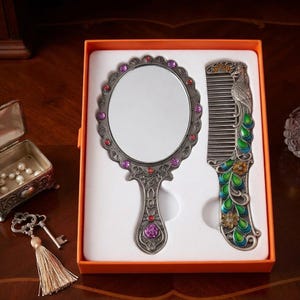 May include: A silver-toned hand mirror and comb set in an orange box. The oval mirror features a scalloped edge with embedded purple and red crystals. The matching comb is decorated with a peacock motif in green, blue, and yellow enamel accents.