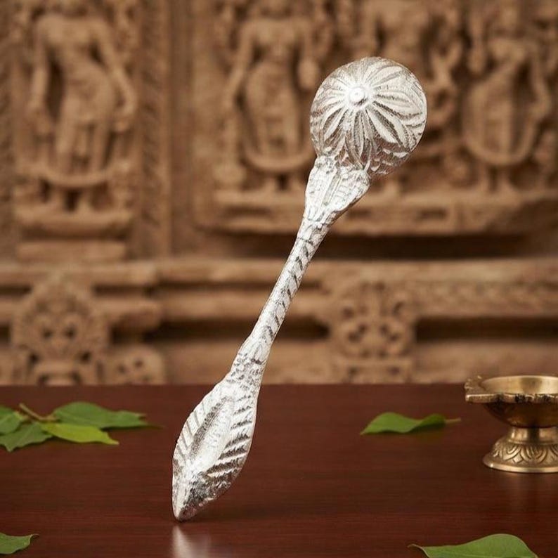 May include: A silver-colored spoon with a round, textured bowl featuring a radial floral pattern. The handle is elongated and tapers to a pointed, leaf-shaped end with intricate, etched detailing along its entire length and surface.