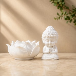 Könnte beinhalten: Eine weiße Keramikfigur eines sitzenden Buddha mit den Händen in Gebetshaltung, platziert neben einer weißen Keramikschale in Form einer Lotusblüte. Beide Gegenstände haben eine glatte, glänzende Oberfläche und sind in reinem Weiß gehalten.