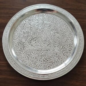 May include: A round silver-colored metal plate featuring an intricate embossed pattern of floral and vine motifs across the center. The rim is decorated with a repeating leaf-like border design, and the surface has a polished, reflective finish.