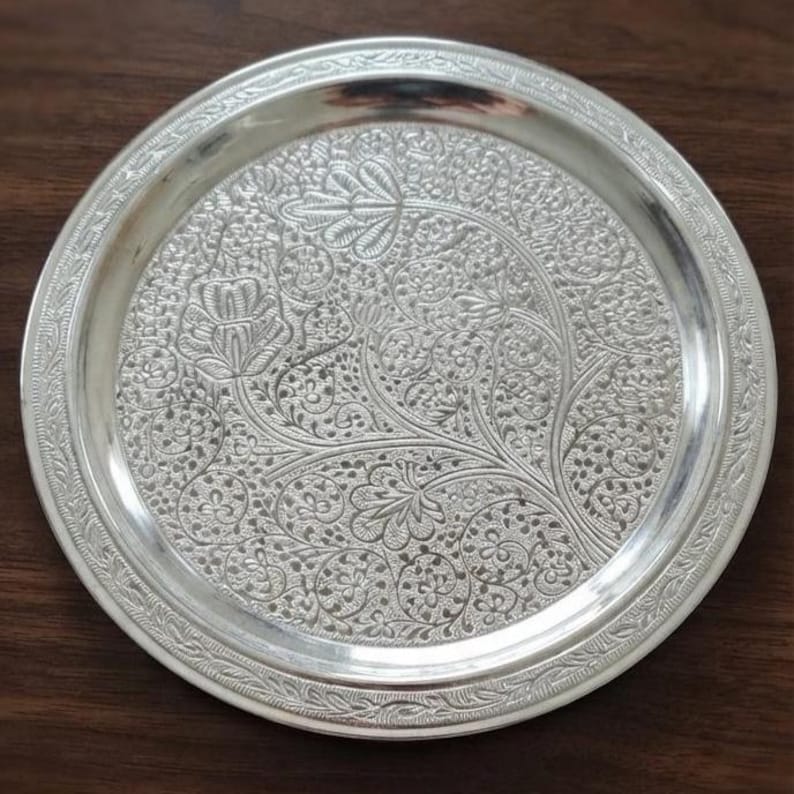 May include: A round silver-colored metal plate featuring an intricate embossed pattern of floral and vine motifs across the center. The rim is decorated with a repeating leaf-like border design, and the surface has a polished, reflective finish.