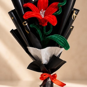 May include: A single red pipe cleaner lily flower with green stems and leaves, wrapped in black paper with small gold heart accents. The bouquet is tied at the base with a red ribbon, featuring a white paper layer beneath the black wrapping.