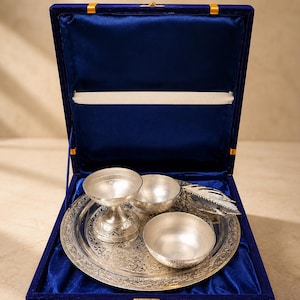 May include: A silver-toned metal set featuring a round engraved tray, two small bowls, a pedestal cup, and a feather-shaped object, all presented inside a rectangular blue velvet-lined display box with a gold-colored metal latch closure.