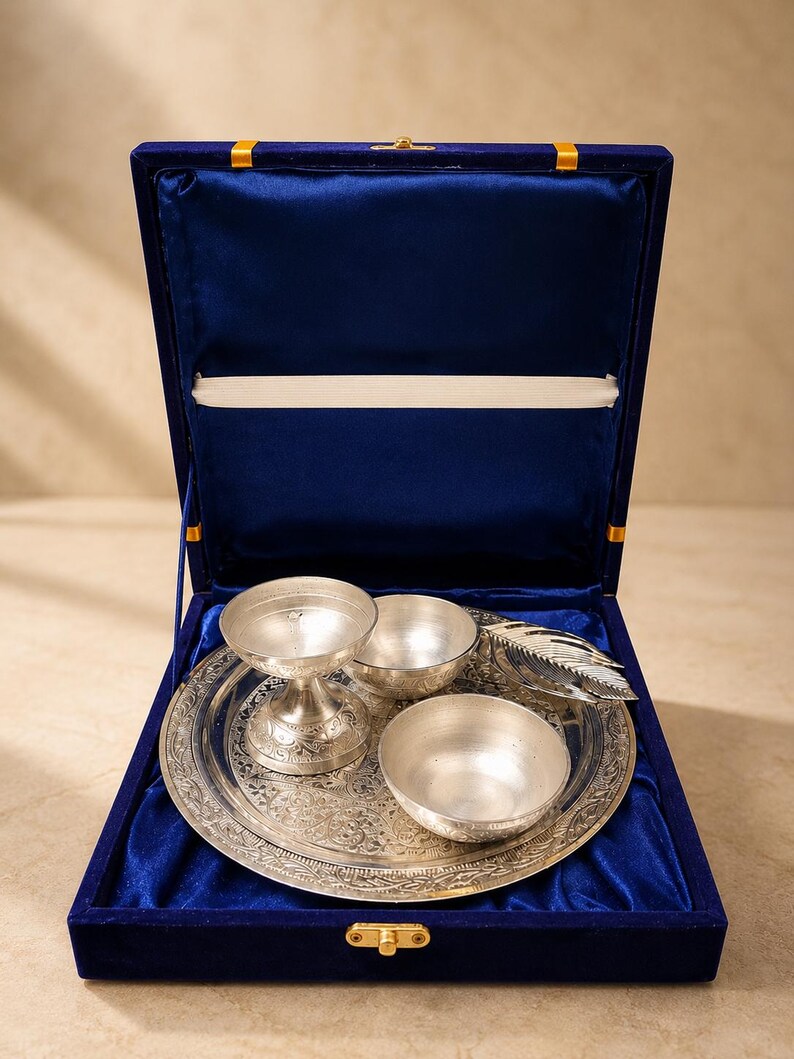 May include: A silver-toned metal set featuring a round engraved tray, two small bowls, a pedestal cup, and a feather-shaped object, all presented inside a rectangular blue velvet-lined display box with a gold-colored metal latch closure.
