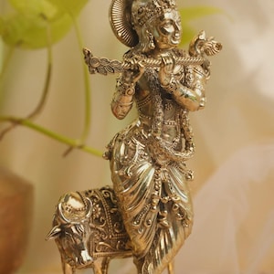 May include: A polished brass statue of a standing figure playing a flute, adorned with intricate jewelry and a crown, positioned beside a small cow. The entire sculpture rests on a circular, textured base with a detailed geometric pattern.