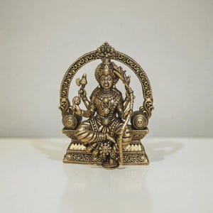 Può includere: Una statuetta in metallo color bronzo raffigurante una divinità con più braccia seduta a gambe incrociate. La figura è incorniciata da un arco circolare ornato e poggia su una base rettangolare, caratterizzata da dettagli in rilievo e motivi.