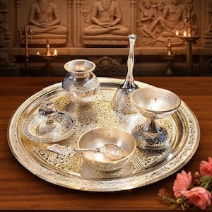 May include: A silver-toned metal puja thali set featuring a large circular tray with intricate floral engravings. The set includes a small pot, a bell with a long handle, a diya lamp, a small bowl with a spoon, and a small plate with a central stand.