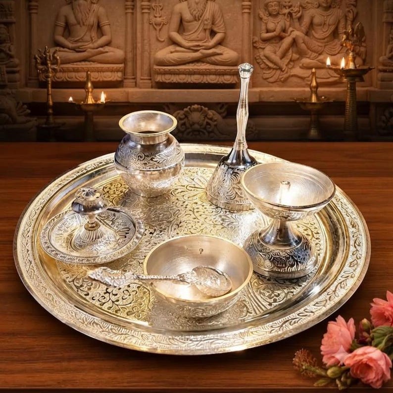 May include: A silver-toned metal puja thali set featuring a large circular tray with intricate floral engravings. The set includes a small pot, a bell with a long handle, a diya lamp, a small bowl with a spoon, and a small plate with a central stand.