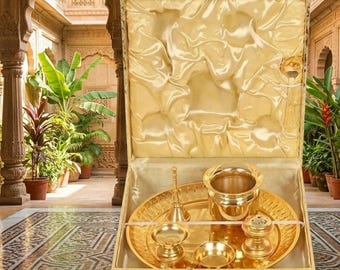 May include: A gold-toned metal ritual set presented in a cream-colored satin-lined box. The set includes a circular tray, a small bowl, a bell, a cylindrical container, and a small dish, all featuring a polished, reflective metallic finish.
