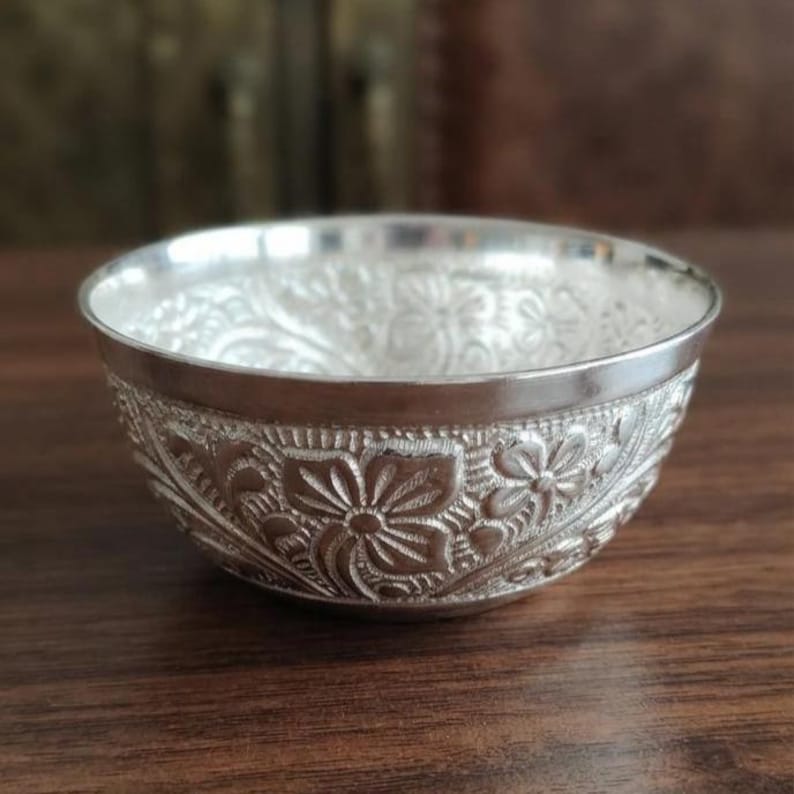 May include: A small, round, silver-colored bowl featuring an intricate, embossed floral pattern covering the entire exterior surface. The bowl has a smooth, polished rim and a deep, rounded interior, showcasing detailed petal and leaf engravings.