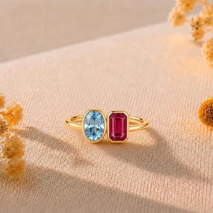 Peut inclure: Une bague en or ornée de deux pierres précieuses distinctes placées côte à côte. L'une est une pierre ovale bleu clair, l'autre est une pierre rectangulaire rose vif taille émeraude. L'anneau est fin et poli, avec un design métallique simple.