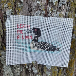 May include: A white needlepoint canvas featuring a cross-stitched black and white loon swimming on light blue water. The text LEAVE ME A LOON is stitched in red capital letters to the left of the bird. The design is on a grid-like fabric.