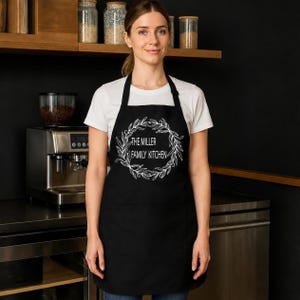 May include: A black apron featuring a white line-art wreath design centred on the chest. Inside the wreath, the text reads THE MILLER FAMILY KITCHEN in a clean, sans-serif font. The apron includes a large front pocket and adjustable neck strap.