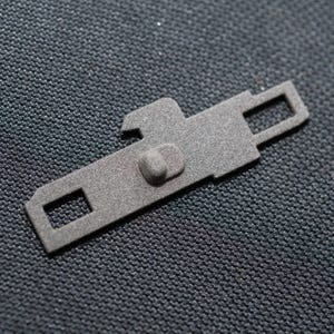 May include: A gray metal zipper pull with a small rectangular opening and a raised button. The zipper pull is designed to be attached to a zipper.