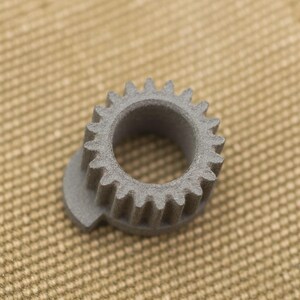 May include: A gray, circular gear with evenly spaced teeth. The gear has a central hole and a small, flat extension on one side. The object is made of a textured material and sits on a tan surface.