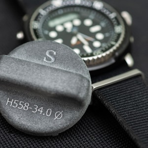 May include: A gray metal cap with the text "S" and "H558-34.0 Ø" engraved on it. The cap is sitting on a black surface next to a black watch with a silver band.