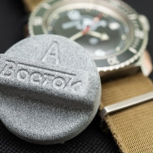 Op de afbeelding: Een grijze metalen horlogekap met het woord "BOSTOK" erop gegraveerd. De dop ligt op een bruine horlogeband.