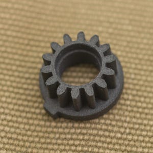 Może przedstawiać: Mała, ciemnoszara zębatka z wieloma zębami. Zębatka ma okrągły środek i jest umieszczona na teksturowanym, beżowym tle. Zęby zębatki są równomiernie rozmieszczone na zewnętrznej krawędzi.