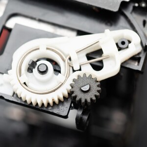 May include: Close-up of mechanical components featuring white and black gears, a spring, and a plastic housing. The gears are interlocked, suggesting a mechanism or machine part. The image is well-lit, highlighting the details of the parts.