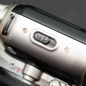 Könnte beinhalten: Ein silberner und schwarzer Camcorder mit einem Knopf, der ein Rückspulsymbol und ein Abspielsymbol hat.