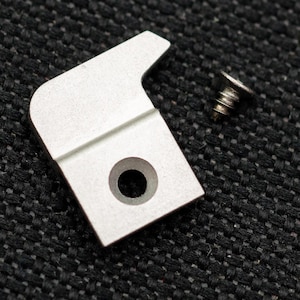 May include: A silver metal hook with a round hole in the center. The hook is on a black textured surface.