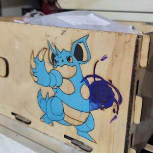 Puede incluir: Una caja de almacenamiento rectangular de madera con un diseño cortado con láser de una criatura azul con cuernos, vientre bronceado y un orbe de energía púrpura oscuro. El lateral de la caja muestra el texto "NIDOKING" grabado en una pequeña placa.