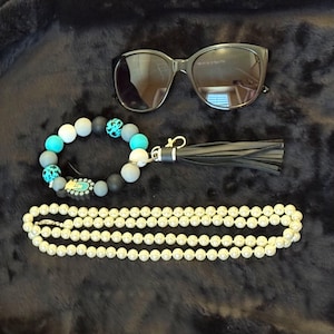 May include: A pair of black sunglasses, a circular keychain featuring matte grey, white, and turquoise beads with a black leather tassel, and a long strand of white faux pearls. The beads include solid colors and turquoise leopard print patterns.