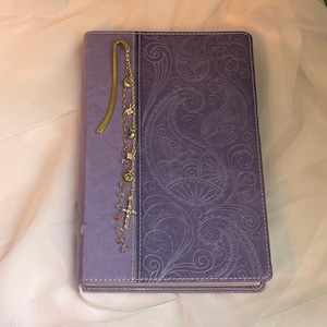 May include: A rectangular purple book with a paisley embossed cover design. A gold-toned metal bookmark is attached to the spine, featuring a long chain adorned with small colorful beads, clear crystals, and a small gold cross charm hanging down.
