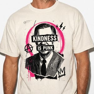 Pode incluir: Uma t-shirt de manga curta cor de creme com um gr&aacute;fico a preto e branco de um homem com um casaco de malha. O design inclui uma pincelada circular rosa, um s&iacute;mbolo de anarquia e o texto "KINDNESS IS PUNK" impresso sobre os olhos.