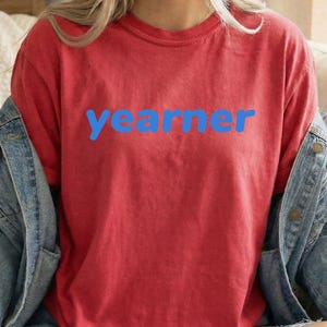 Op de afbeelding: Een rood T-shirt met korte mouwen en het woord "yearner" in een vetgedrukt, blauw schreefloos lettertype op de borst, gedragen onder een blauw spijkerjack met zichtbare metalen knopen en een klassieke kraag.
