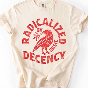 May include: A cream-colored short-sleeve t-shirt featuring a red graphic print. The design displays a bird holding a small branch, surrounded by the text "RADICALIZED BY BASIC DECENCY" in a circular, distressed, bold red font style.