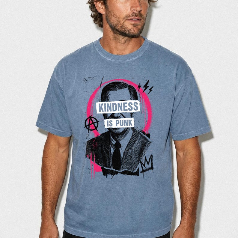 Pode incluir: Uma t-shirt azul de manga curta e gola redonda com uma ilustra&ccedil;&atilde;o gr&aacute;fica a preto e branco de um homem com um casaco de malha e gravata. O design inclui um c&iacute;rculo cor-de-rosa, um s&iacute;mbolo anarquista e o texto "KINDNESS IS PUNK".