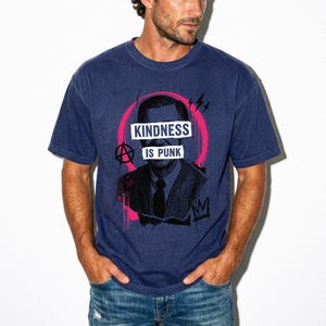 Pode incluir: Uma t-shirt azul-marinho de manga curta com uma estampa gr&aacute;fica de uma pessoa de fato e gravata. O design inclui um c&iacute;rculo rosa brilhante, um s&iacute;mbolo de anarquia e o texto branco "KINDNESS IS PUNK" sobre o centro do rosto.