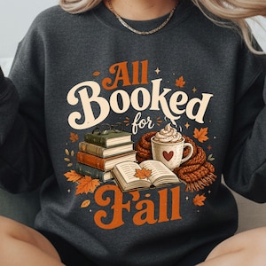 May include: A dark charcoal gray crewneck sweatshirt featuring a graphic print with the text All Booked for Fall. The design includes a stack of books, an open book, a mug with whipped cream and a heart, a scarf, and scattered autumn leaves.