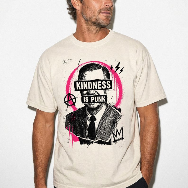 Pode incluir: Uma t-shirt de manga curta cor de creme com um gr&aacute;fico a preto e branco de um homem com um casaco de malha. O design inclui uma pincelada circular rosa, um s&iacute;mbolo de anarquia e o texto "KINDNESS IS PUNK" impresso sobre os olhos.
