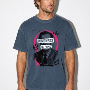 Pode incluir: Uma t-shirt de manga curta azul ard&oacute;sia com um gr&aacute;fico a preto e branco de uma pessoa com um casaco de malha e gravata. O design inclui um detalhe circular cor-de-rosa vivo, um s&iacute;mbolo anarquista e o texto "KINDNESS IS PUNK" em letras brancas.