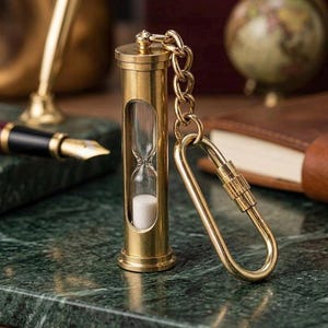 May include: A brass hourglass keychain featuring a cylindrical metal frame with a clear glass center containing white sand. The hourglass is attached to a sturdy metal chain and a screw-lock carabiner, providing a functional and metallic accessory.