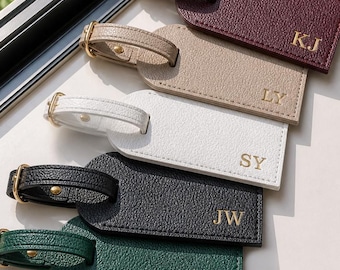 May include: Five rectangular luggage tags with a textured finish, each featuring a gold-toned buckle strap and gold-embossed initials. The colors include burgundy with KJ, beige with LY, white with SY, black with JW, and dark green with RC.